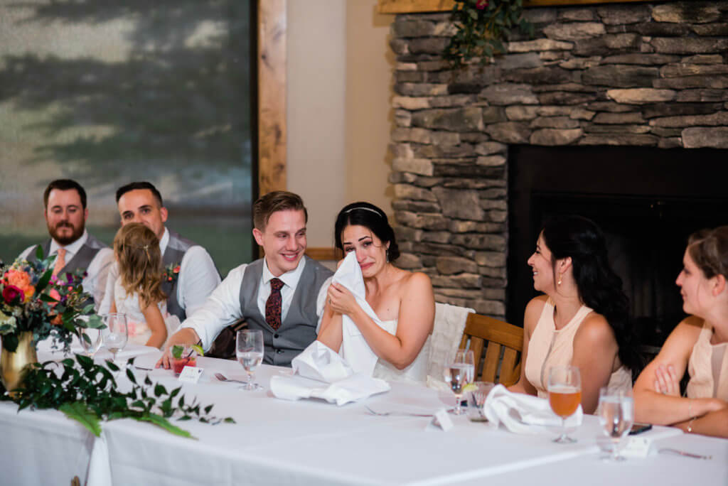 A bride wipes happy tears from her eyes sitting next to her new husband at the wedding table.